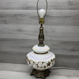 May include: A vintage table lamp with a white, iridescent glass base and a brass-colored metal stem and base. The base features gold-colored floral and star designs. The lamp has a white lightbulb and a curved metal arm.