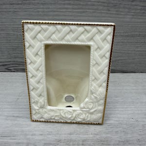 May include: A rectangular cream-coloured ceramic picture frame featuring a raised basket-weave pattern on the border and sculpted rose floral details at the bottom corners. The outer edge is finished with a thin, metallic gold-toned trim.