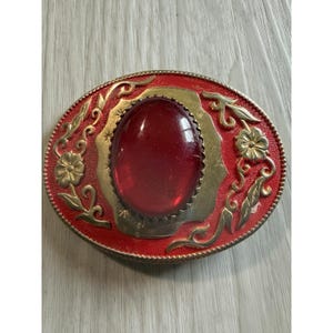 Puede incluir: Una hebilla de cinturón ovalada con una gran piedra cabujón roja y lisa en el centro, engastada en un marco de metal dorado. El fondo rojo texturizado está decorado con motivos florales y de enredaderas en relieve de color dorado.