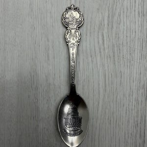 May include: A silver spoon with a detailed design. The spoon features an image of the U.S. Capitol building and the text "WASHINGTON DC". The handle has the text "DIST. OF COLUM." and an ornate top with a landscape scene.