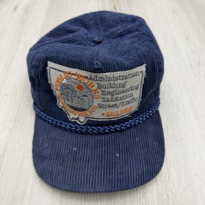 May include: A navy blue corduroy baseball cap featuring a braided trim above the brim and a rectangular patch. The patch displays the text Public Works Administration, Building, Engineering, Sanitation, Street/Traffic, Working For You, and Billings.