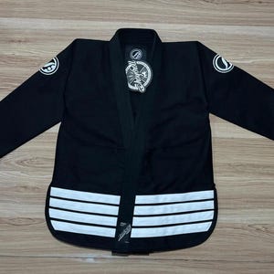 May include: Black Jiu-Jitsu uniform with white accents. The uniform features a black jacket with white stripes along the bottom and circular patches on the sleeves. A tag hangs from the collar. The uniform is laid out on a wooden surface.