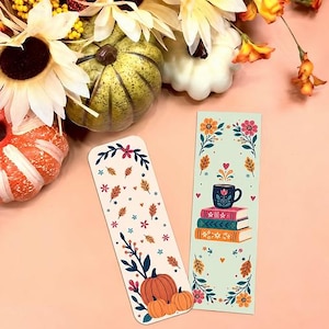 May include: Two rectangular bookmarks with rounded corners. One bookmark features a white background with scattered autumn leaves, small flowers, and three orange pumpkins at the base. The other bookmark has a light green background with a stack of three books, a mug, and floral motifs.