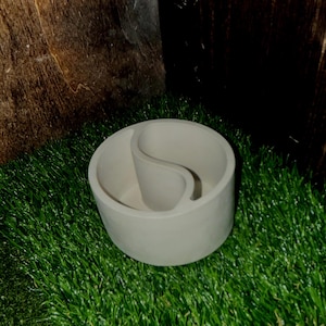 May include: A circular, light beige container featuring an internal divider shaped like a yin-yang symbol. The object has smooth, solid walls and a flat base, creating two distinct, curved compartments within the single cylindrical structure.