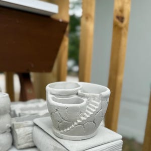 Op de afbeelding: Een lichtgrijze betonnen plantenbak met een ontwerp op meerdere niveaus, twee afzonderlijke ronde openingen en een kleine trap die in de buitenwand is gegoten. Het oppervlak heeft een getextureerde, gebarsten steenlook.