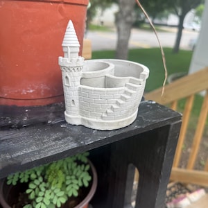 May include: A light grey concrete desk organiser shaped like a castle, featuring a tall turret, a brick-patterned wall, and a set of stairs leading up to the rim. The interior is divided into two circular compartments for holding small items.