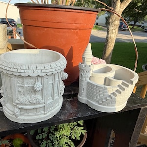 Peut inclure: Deux jardinières en béton blanc cassé. La jardinière de gauche est cylindrique avec un relief représentant une devanture de magasin et des paniers de fleurs suspendus. Celle de droite a la forme d'un petit château avec une tour et un escalier.