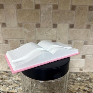 May include: A white, open book-shaped object with a pink border sits atop a black lid. The book is 3D-printed and rests on a clear glass jar with a textured pattern. The background features a neutral-toned tile backsplash.
