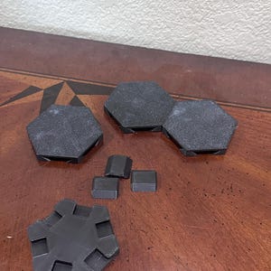 May include: A collection of black, geometric 3D-printed objects. The set includes several hexagonal and square shapes, with a textured surface. The objects are arranged on a wooden surface.