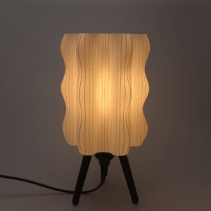 May include: A cream-colored table lamp with a wavy, textured shade and a black tripod base. The lamp is lit, emitting a warm glow. The shade has vertical lines, and the lamp is plugged into a wall outlet.