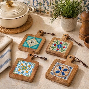 May include: Four wooden cutting board style coasters with colorful, patterned tile inserts. Each coaster has a handle and a leather loop. The coasters are arranged on a light-colored surface, with a white pot and a small plant in the background.