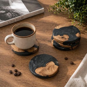 Puede incluir: Juego de posavasos redondos con un diseño único, combinando madera y resina negra. Una taza de café descansa sobre uno de ellos, con una pila de posavasos cerca. Granos de café dispersos. Cada posavasos mide aproximadamente 10 cm de diámetro.