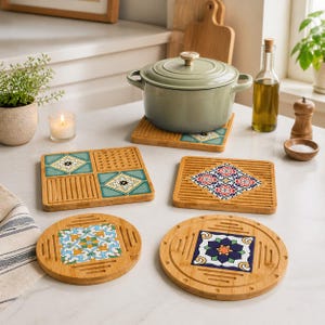 May include: A collection of bamboo trivets with colorful, patterned ceramic tile inserts. The trivets are in various shapes, including square and round, and feature floral and geometric designs. A green pot sits on one of the trivets.