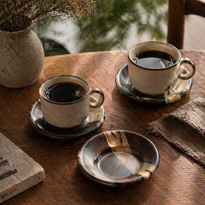 以下が含まれることがあります： 濃いコーヒーが入った3つのセラミックコーヒーカップ。それぞれにマッチするソーサーが付いています。カップとソーサーは、茶色のトリムが付いた、テクスチャード加工された斑点のあるベージュ仕上げです。空のソーサーも1つ見えます。シーンは木製のテーブルに設定されています。