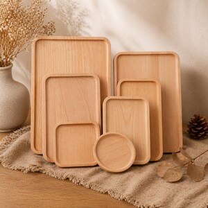 May include: A collection of light-colored wooden serving trays in various rectangular and square shapes. The trays are stacked together, showcasing different sizes, with a round coaster in the front. The wood grain is visible.