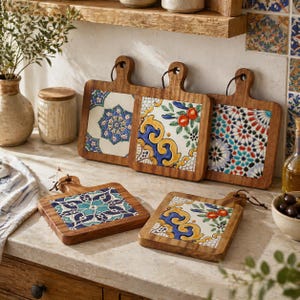 May include: Wooden cutting boards with decorative ceramic tile inserts. The tiles feature colorful patterns in blue, yellow, and red. Each board has a handle with a leather loop for hanging. The boards are displayed on a countertop.