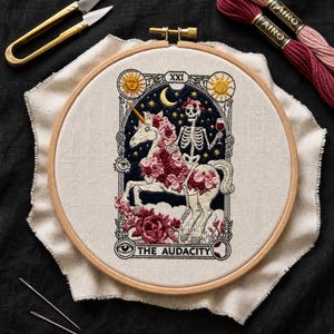 Può includere: Un telaio da ricamo contenente un pannello in tessuto con un disegno ricamato di uno scheletro che cavalca un unicorno decorato con rose rosa e rosse. Lo scheletro tiene in mano un bicchiere di vino rosso. Il testo THE AUDACITY è ricamato in basso.