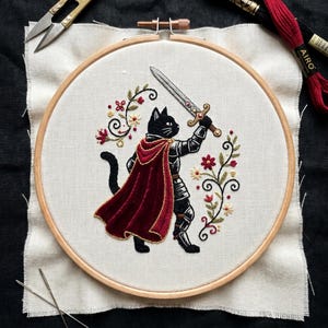 May include: An embroidery hoop containing a piece of off-white fabric with a hand-stitched design of a black cat wearing silver armour and a deep red velvet-textured cape, holding a silver sword, surrounded by decorative floral vine patterns.
