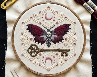 May include: A circular wooden embroidery hoop containing a fabric piece with a hand-stitched design of a moth with a skull pattern on its thorax, a large ornate key, two crescent moons, and small floral accents in shades of burgundy and cream.