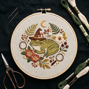 May include: A circular wooden embroidery hoop containing a fabric panel with a stitched green frog wearing a brown hat with small mushrooms. The design includes a crescent moon, ferns, flowers, berries, and two magnifying glasses. Embroidery needles, scissors, and skeins of green and cream thread are arranged around the hoop.