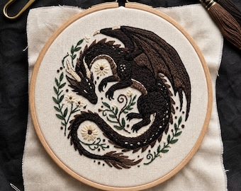 May include: A circular wooden embroidery hoop containing a fabric piece with a hand-stitched dark brown dragon curled among green leaves and small white flowers. The design is centered on off-white fabric, accompanied by gold thread snips and needles.