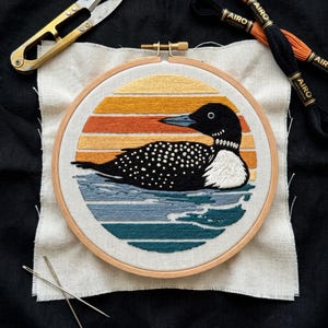 May include: An embroidery hoop containing a stitched loon on water against a sunset background. The loon is black with white spots and a white neck patch. The sunset features horizontal bands of yellow and orange, while the water is blue.