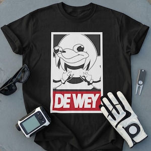 May include: A black short-sleeved t-shirt featuring a white graphic of a stylised cartoon character inside a rectangular frame, with the text DE WEY printed in white on a red background below the character. The shirt is surrounded by golf accessories.
