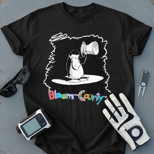 May include: A black short-sleeved t-shirt featuring a white graphic of a penguin tipping its top hat. Below the graphic, the text Bloom County is printed in colorful, stylized letters. The shirt is surrounded by sunglasses, a golf glove, and a divot tool.