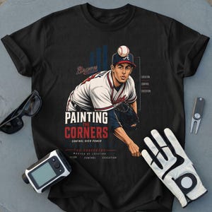 Może przedstawiać: Czarny t-shirt z grafiką miotacza baseballowego w białym mundurze i czerwono-niebieskiej czapce. Tekst głosi PAINTING THE CORNERS, CONTROL OVER POWER, THE PROFESSOR, MASTER OF LOCATION, CONTROL, EXECUTION oraz Braves.