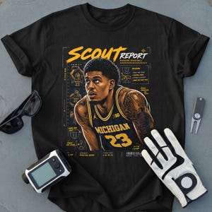 May include: A black t-shirt featuring a graphic of a basketball player in a navy and yellow Michigan jersey with the number 23. The design includes the text SCOUT REPORT, basketball play diagrams, and technical notes in yellow and white ink.