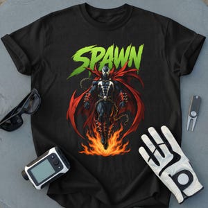 Puede incluir: Una camiseta negra con un gráfico del personaje Spawn con capa roja, cadenas y llamas en los pies. La palabra SPAWN está impresa en letras verdes grandes sobre el personaje. Unas gafas negras, un guante blanco y una herramienta están cerca.