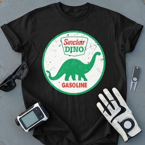 May include: A black short-sleeved t-shirt featuring a circular graphic with a distressed white background and a green border. The graphic displays a green dinosaur silhouette, the text Sinclair DINO, and the word GASOLINE in red lettering.
