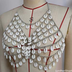 Puede incluir: Un top de malla plateada adornado con numerosas conchas blancas. El top de estilo halter presenta múltiples filas de cadenas, cada una suspendiendo las conchas. Las conchas son ovaladas y están espaciadas uniformemente.