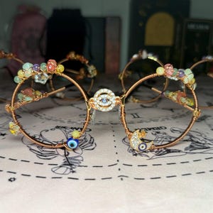 Puede incluir: Monturas de gafas envueltas en alambre de cobre adornadas con piedras preciosas de colores y abalorios decorativos. Las monturas presentan un adorno central en forma de diamante y un pequeño amuleto de ojo azul. Otras monturas son visibles en el fondo.