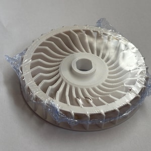 Peut inclure: Une roue de ventilateur circulaire blanche dotée de nombreuses pales incurvées rayonnant à partir d'un moyeu central avec une ouverture hexagonale. Le composant est partiellement enveloppé dans un film plastique transparent et fin.