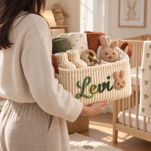 May include: A cream-coloured corduroy storage basket featuring the name Levi embroidered in green script and a small plush bunny face. The basket contains baby items including knit booties, muslin cloths, a wooden teether, and a plush bunny toy.