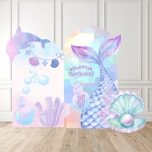 May include: A mermaid-themed birthday party backdrop featuring a dolphin, bubbles, and a mermaid tail. The backdrop includes the text "Happy Birthday Emma" and the number "5". A large pearl in an open shell is also included.
