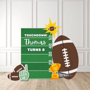 May include: A football-themed birthday party decoration set. Includes a green field backdrop with the text "TOUCHDOWN! Thomas TURNS 8", footballs, a foam finger, a trophy, and a pennant that says "FOOTBALL".