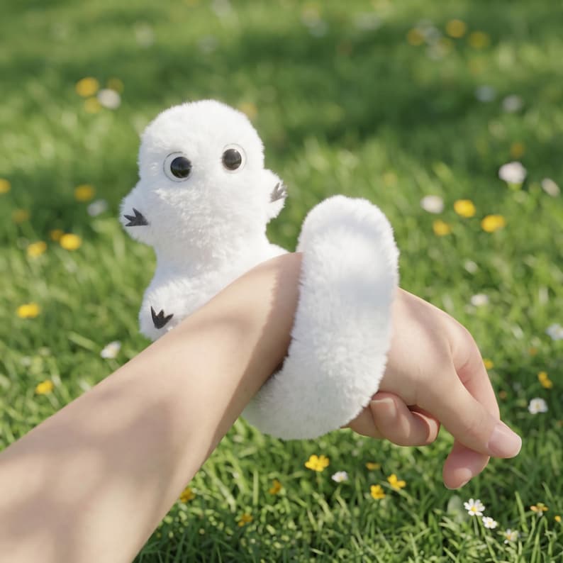 May include: A white plush toy shaped like a small creature with large black eyes and small black feet. The toy is held in place by a white, fluffy wristband. The background is a blurred green field with small yellow and white flowers.