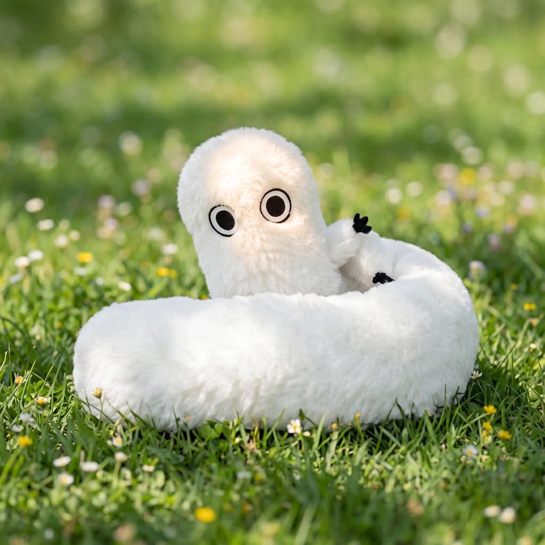 May include: A fluffy white toy, resembling a worm or caterpillar, with big black and white eyes. It rests on a bed of green grass, dotted with small white and yellow flowers.