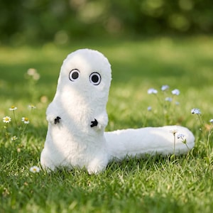 May include: A white plush toy in the shape of a creature with large, round eyes and small black hands. The toy has a long, fluffy tail and is on green grass with small white and blue flowers.