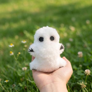 May include: A small, white plush toy with large, black eyes and tiny black claws. The soft, fluffy creature is held in a hand against a backdrop of green grass and wildflowers. The toy has a rounded body and small arms.