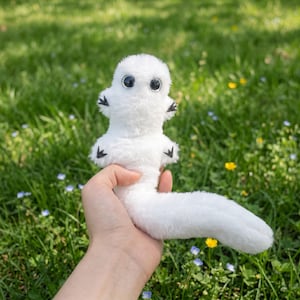 May include: A white plush toy creature with large black eyes and small black accents on its arms. The toy has a long, curved tail and is held against a backdrop of green grass and wildflowers.