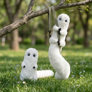 May include: Two white plush toys with black eyes and small black appendages. One toy is hanging from a green strap attached to a tree branch, while the other stands on the grass. The toys have a soft, fuzzy texture and are set against a backdrop of green grass and trees.