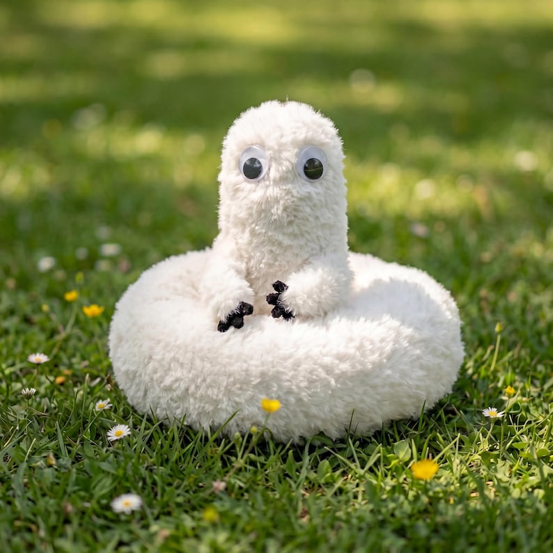 May include: A fluffy white soft toy with large googly eyes and tiny black paws. The plush creature is sitting on green grass with small yellow and white flowers.