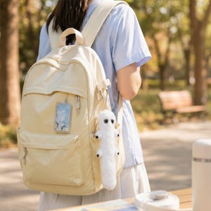 May include: A light beige backpack with multiple pockets and a top handle. A white, fuzzy, worm-like keychain hangs from the zipper. A clear case with a landscape image is also attached. The backpack is worn over the shoulders.