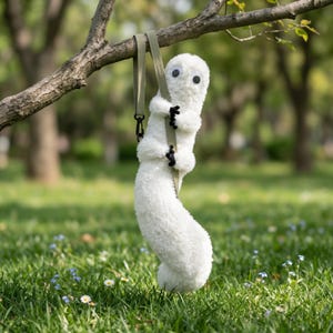 May include: A fluffy white toy with large googly eyes and black claws hangs from a green strap attached to a tree branch. The toy has a long, curved body against a backdrop of green grass and trees.