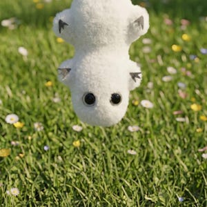 May include: A white plush toy with large black eyes and small gray ears is suspended upside down against a backdrop of green grass and wildflowers. The toy has a soft, fluffy texture.