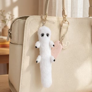 May include: A white plush keychain in the shape of a creature with black paws and eyes. The keychain is attached to a light pink tag and a silver chain, hanging from a cream-colored handbag.