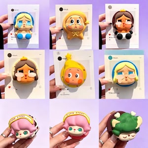 Puede incluir: Una colección de coloridos agarres para teléfono de estilo caricatura. Cada agarre presenta un diseño de personaje único con caras expresivas y colores vibrantes, incluyendo amarillo, rosa y azul. Los agarres están montados en tarjetas con marcos de estilo redes sociales.